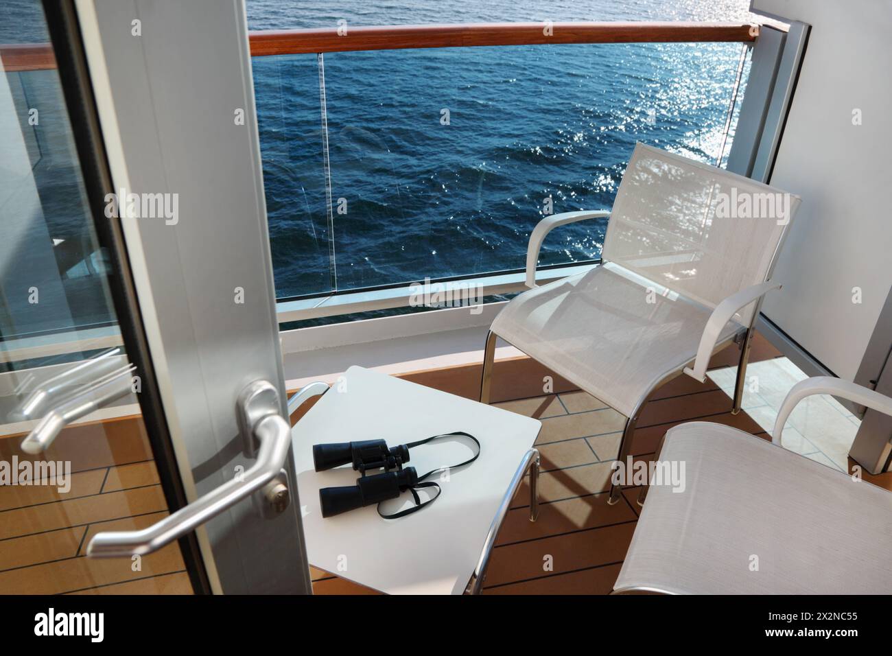 open door; glassy balcony with white chairs and table with binoculars on cruise liner deck Stock Photo