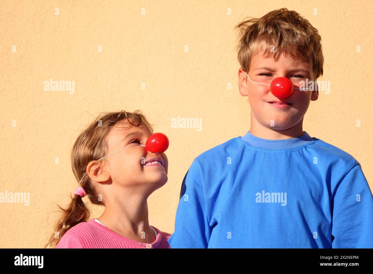 Brother and sister with red clown noses stand near wall and smiles. Girl looks at boy. Stock Photo