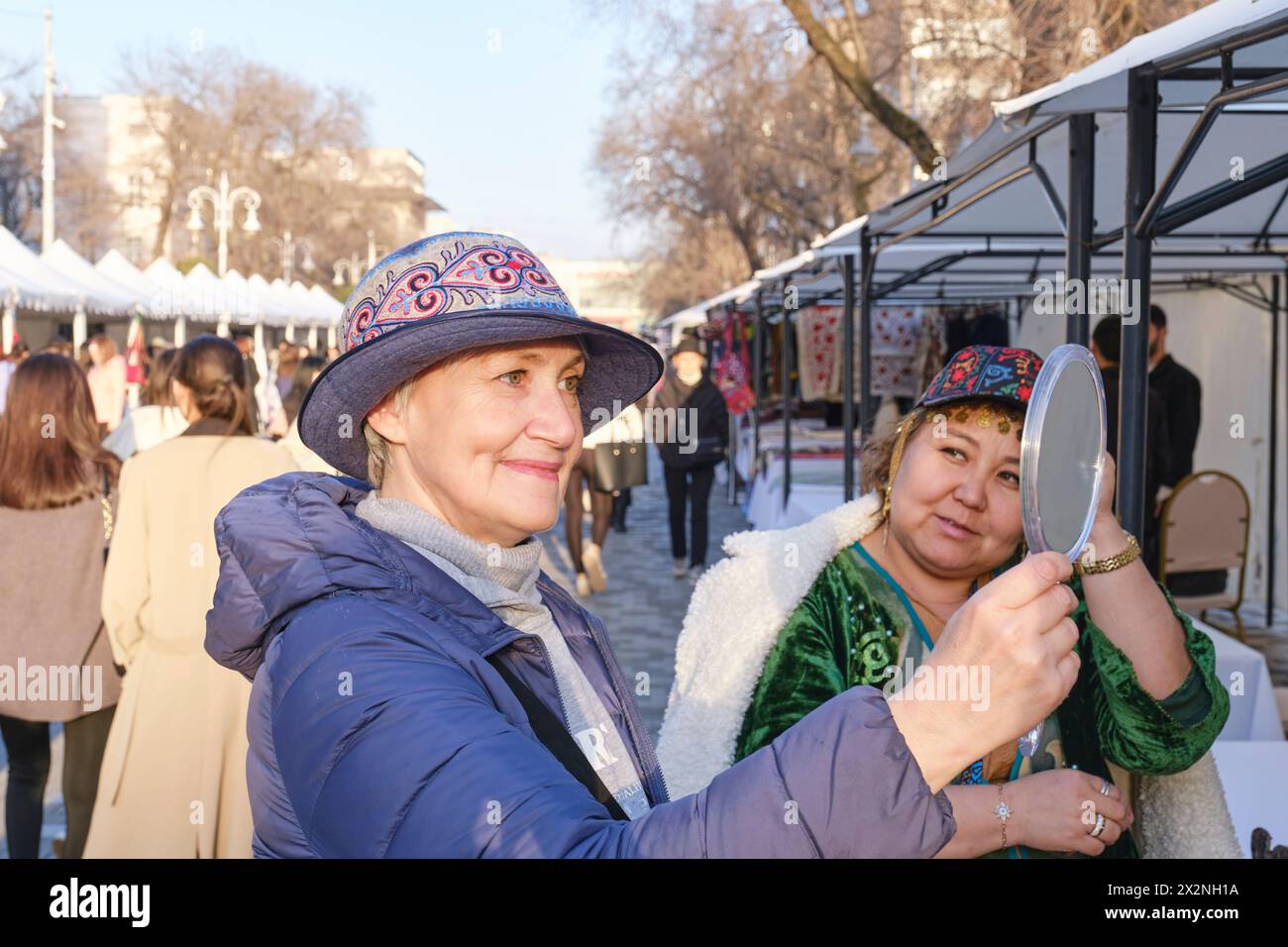 Almaty, Kazakhstan - March 18, 2024: Senior Caucasian woman trying on a felt hat with a traditional Kazakh pattern, looking in the mirror near a sales Stock Photo