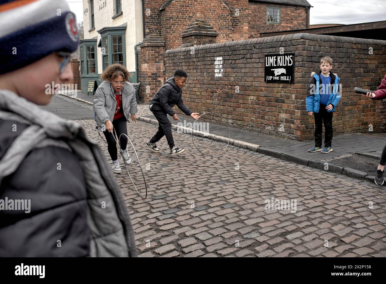 Children rolling a metal hoop and stick on the cobbled streets at the Black Country Museum. 1900's child's toy. England UK Stock Photo