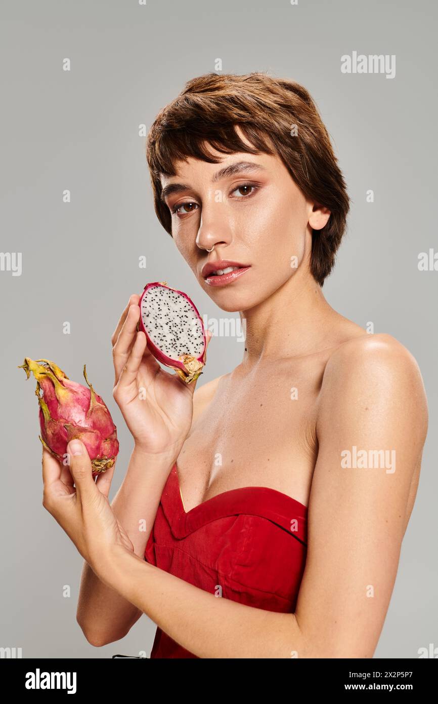 A young woman in a red dress holding a dragon fruit, surrounded by vibrant tropical fruits. Stock Photo