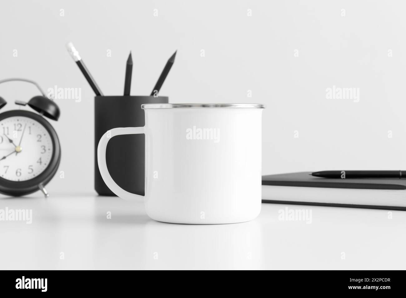 Enamel mug mockup with workspace accessories on a white table. Stock Photo