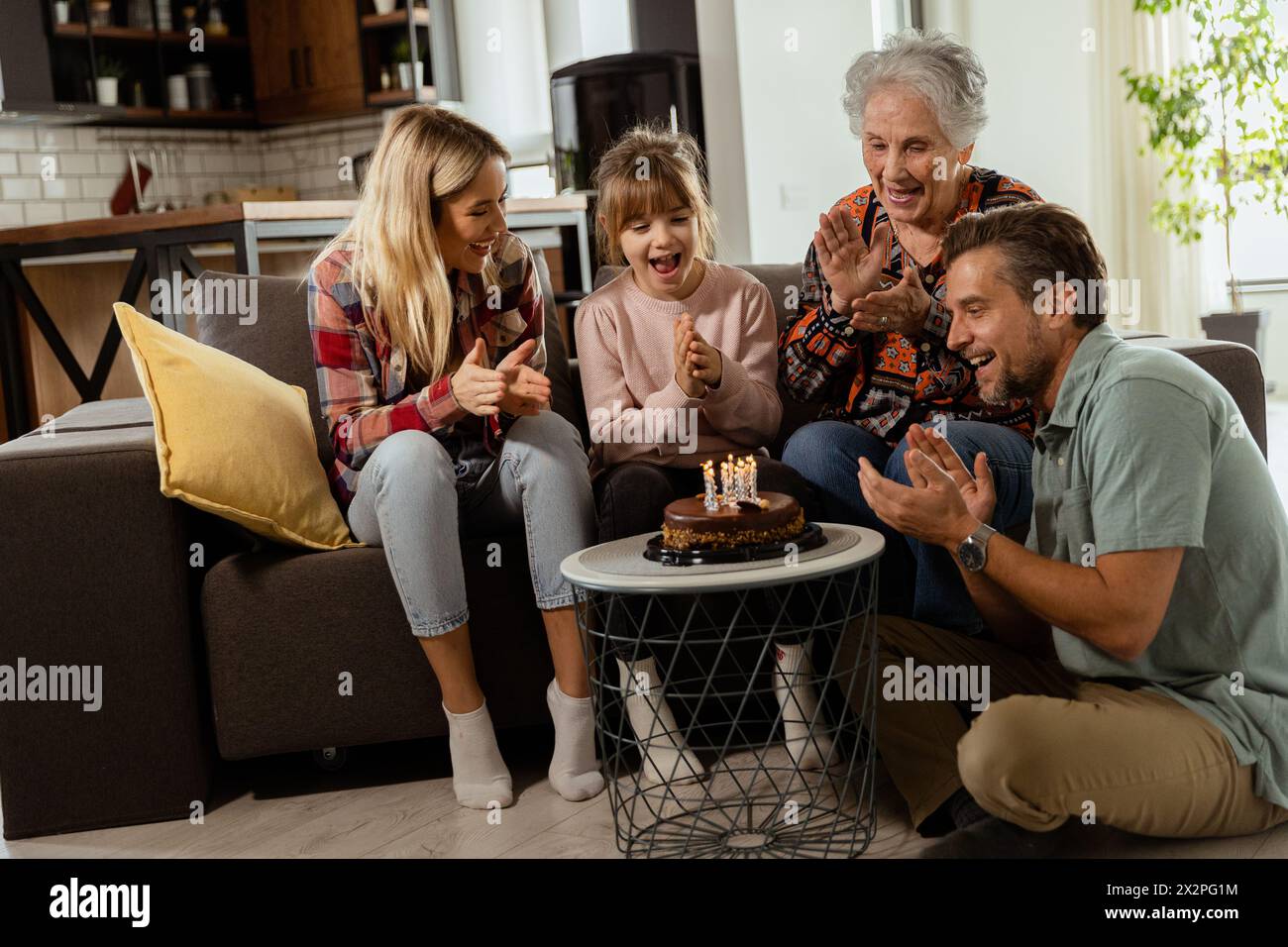 Heartwarming scene unfolds as a multi-generational family gathers on a couch to present a birthday cake to a delighted grandmother, creating memories Stock Photo