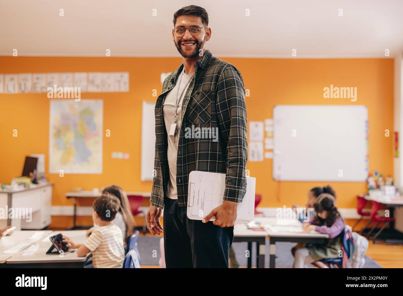 Happy elementary school teacher standing in a classroom, with children working at their desks in the background. Man teaching kids in an early child d Stock Photo