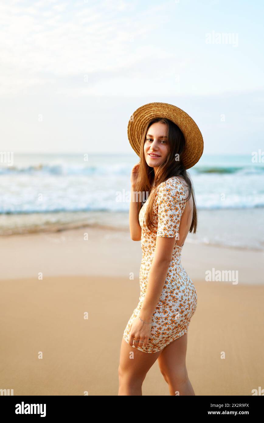 Smiling woman in patterned summer dress, straw stands on sandy beach at  sunset. Relaxed tourist enjoys evening sea breeze. Golden hour sunlight  Stock Photo - Alamy