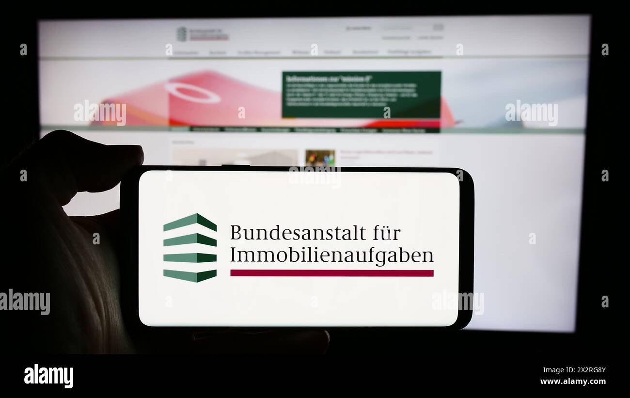 Person holding cellphone with logo of German agency Bundesanstalt für Immobilienaufgaben (BImA) in front of webpage. Focus on phone display. Stock Photo