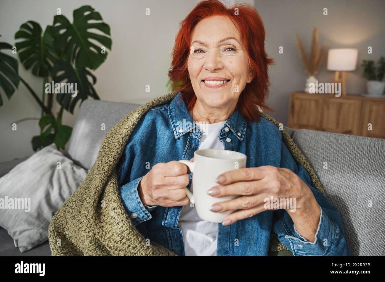Smiling senior woman sitting under blanket on sofa drinking tea at home Stock Photo