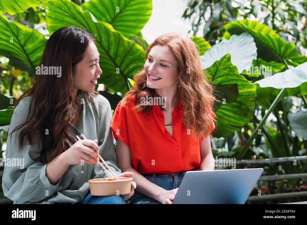 Smiling woman eating meal and talking with friend near plants Stock Photo