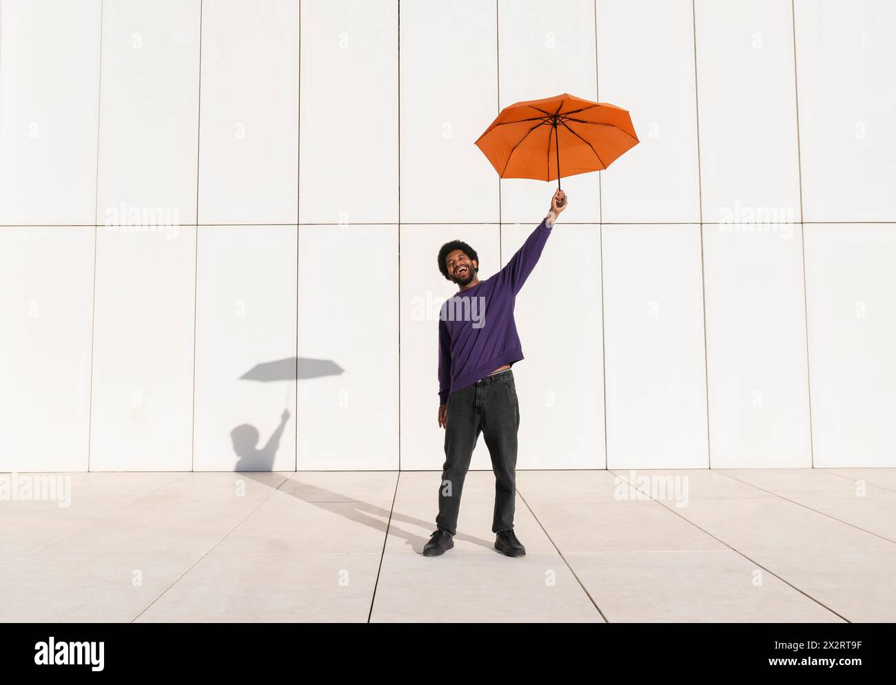 Happy young man standing with orange umbrella in front of white wall ...