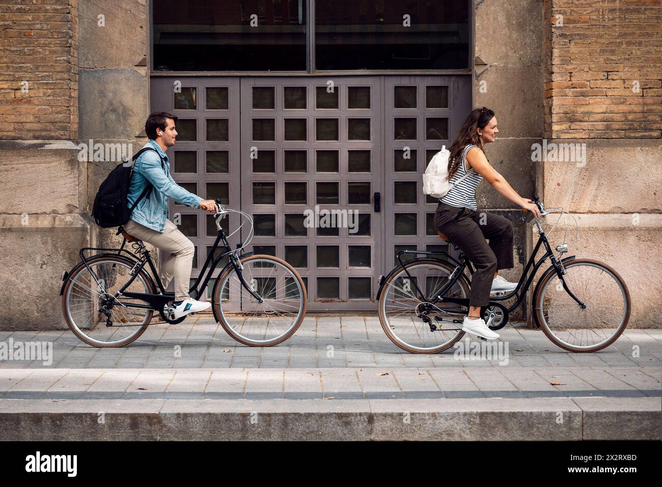 Couple cycling near building on sidewalk Stock Photo