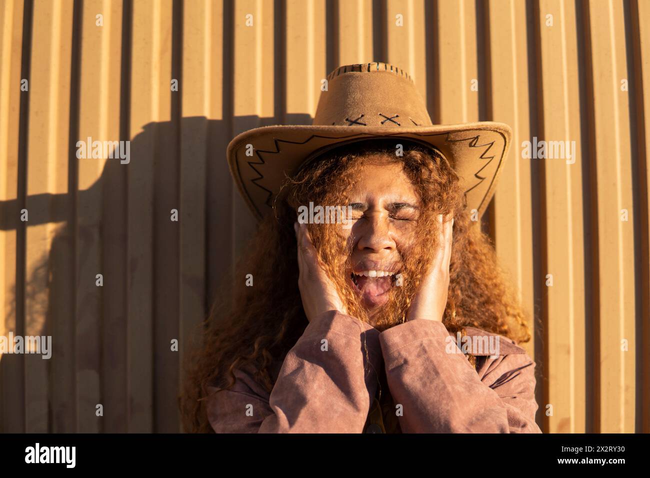 Curly haired woman wearing cowboy hat shouting in front of orange metal wall Stock Photo
