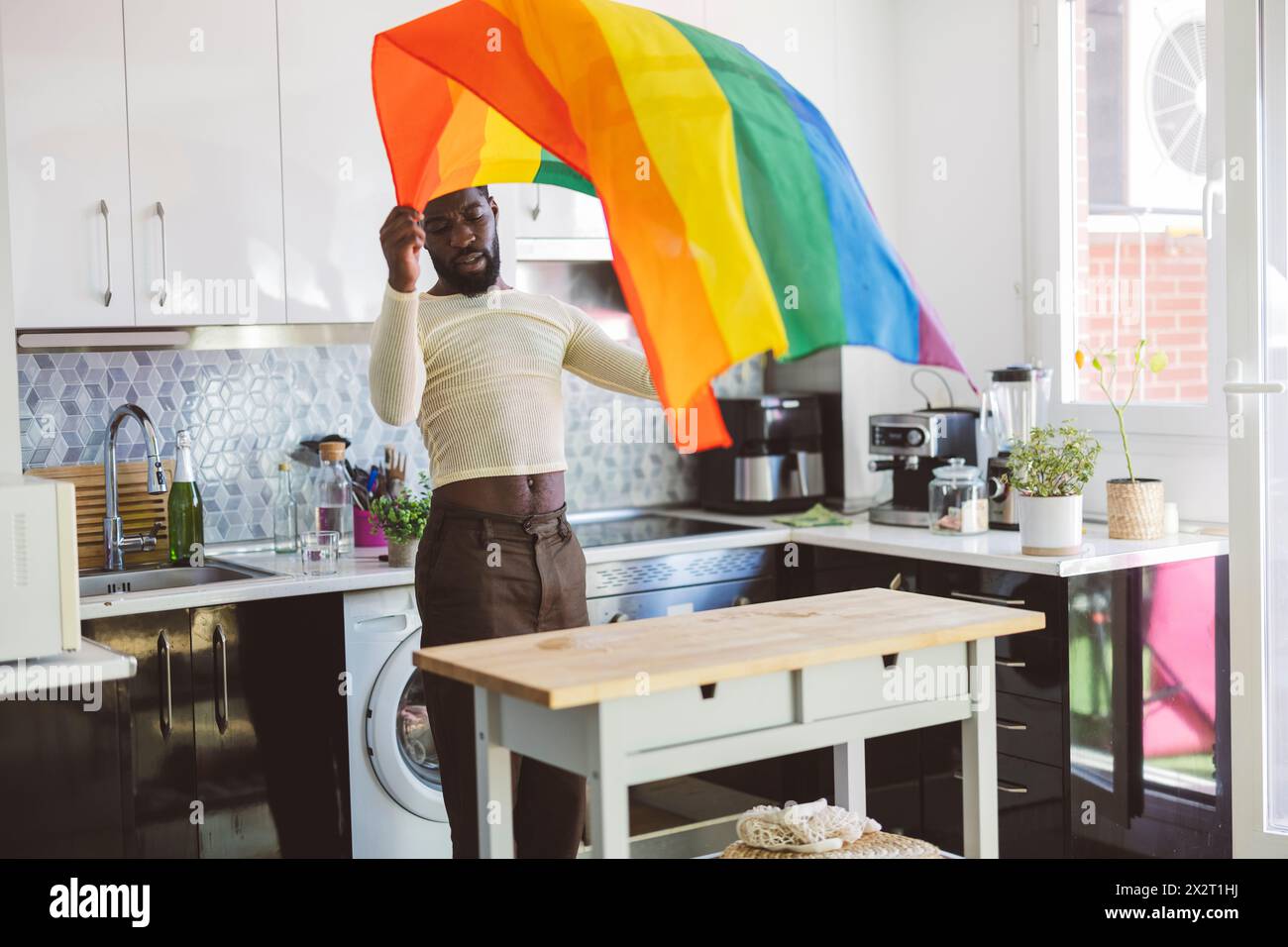 Young non-binary person holding rainbow flag near table at home Stock Photo