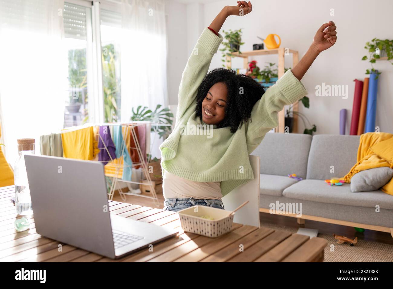 Happy young businesswoman stretching hands sitting with laptop and meal at table in living room Stock Photo
