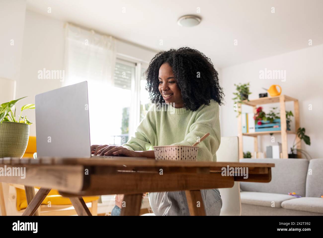 Happy young businesswoman using laptop sitting with meal at table in living room Stock Photo