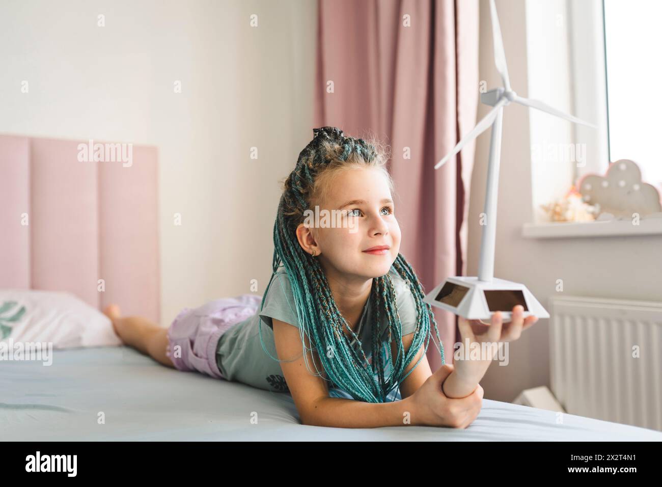 Smiling girl with dyed braided hair holding wind turbine model lying on bed at home Stock Photo