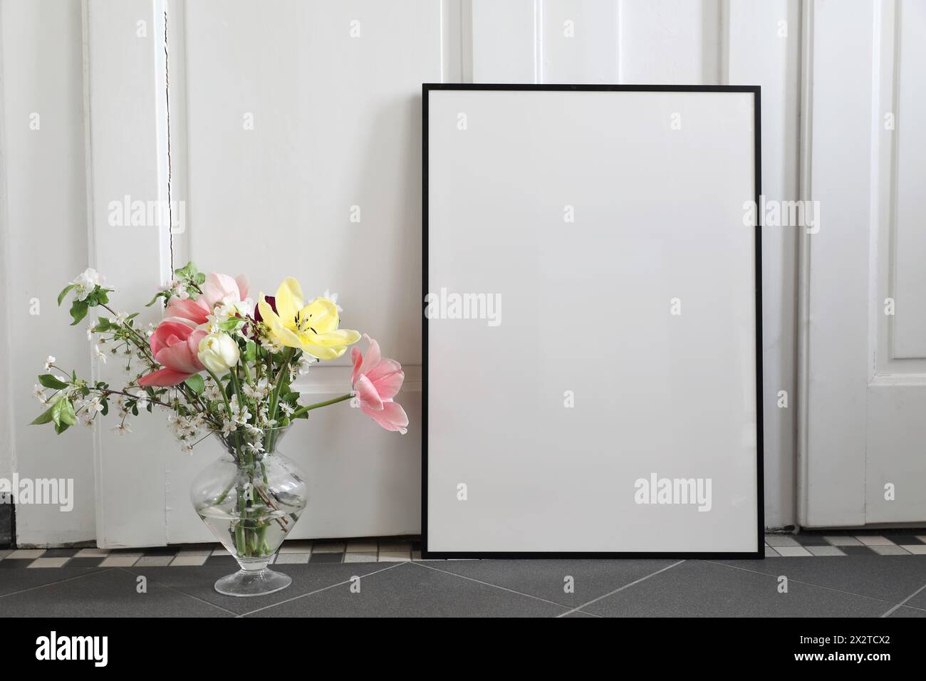 Spring still life scene. Tulips, cherry tree blossoms bouquet in glass vase. Blank black picture frame, poster mockup on grey tile floor. White wall Stock Photo
