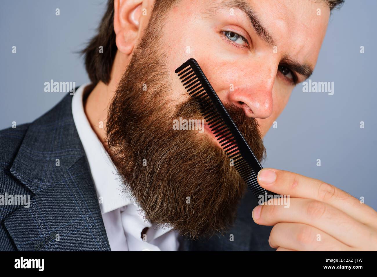 Barbershop. Stylish bearded man in suit combing beard with haircomb. Portrait of brutal man with beard and mustache with barber comb. Professional Stock Photo