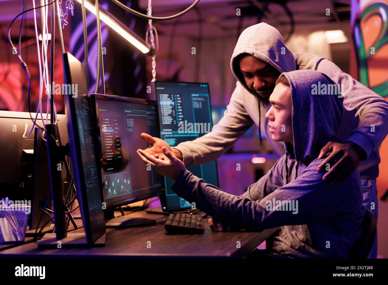 Two hackers collaborating while breaching data and coding malicious software on computer. Diverse criminals working together while programming ransomware in abandoned warehouse Stock Photo