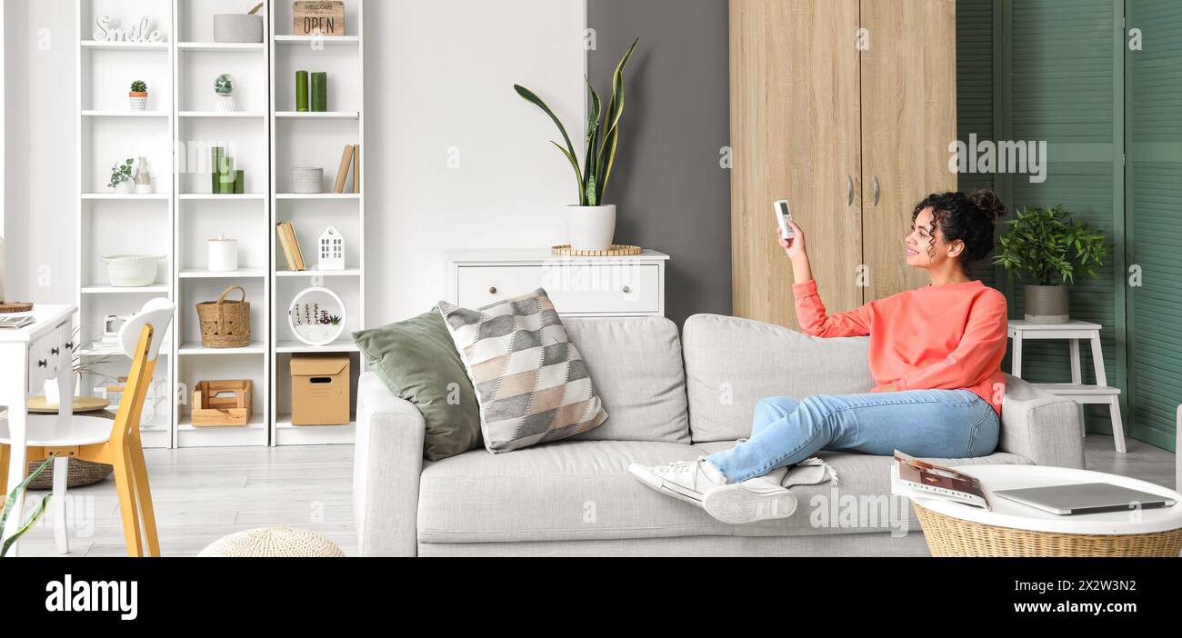 Young woman switching on air conditioner while sitting on sofa at home Stock Photo