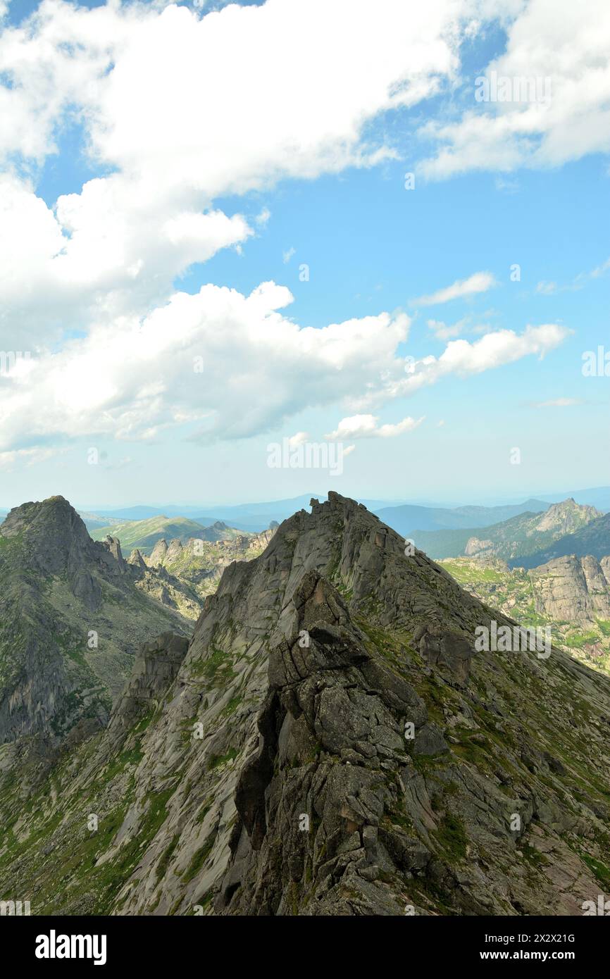A narrow ridge of high rock with steep peaks in a chain of picturesque mountains under a cloudy sky on a clear summer day. Ergaki Natural Park, Krasno Stock Photo