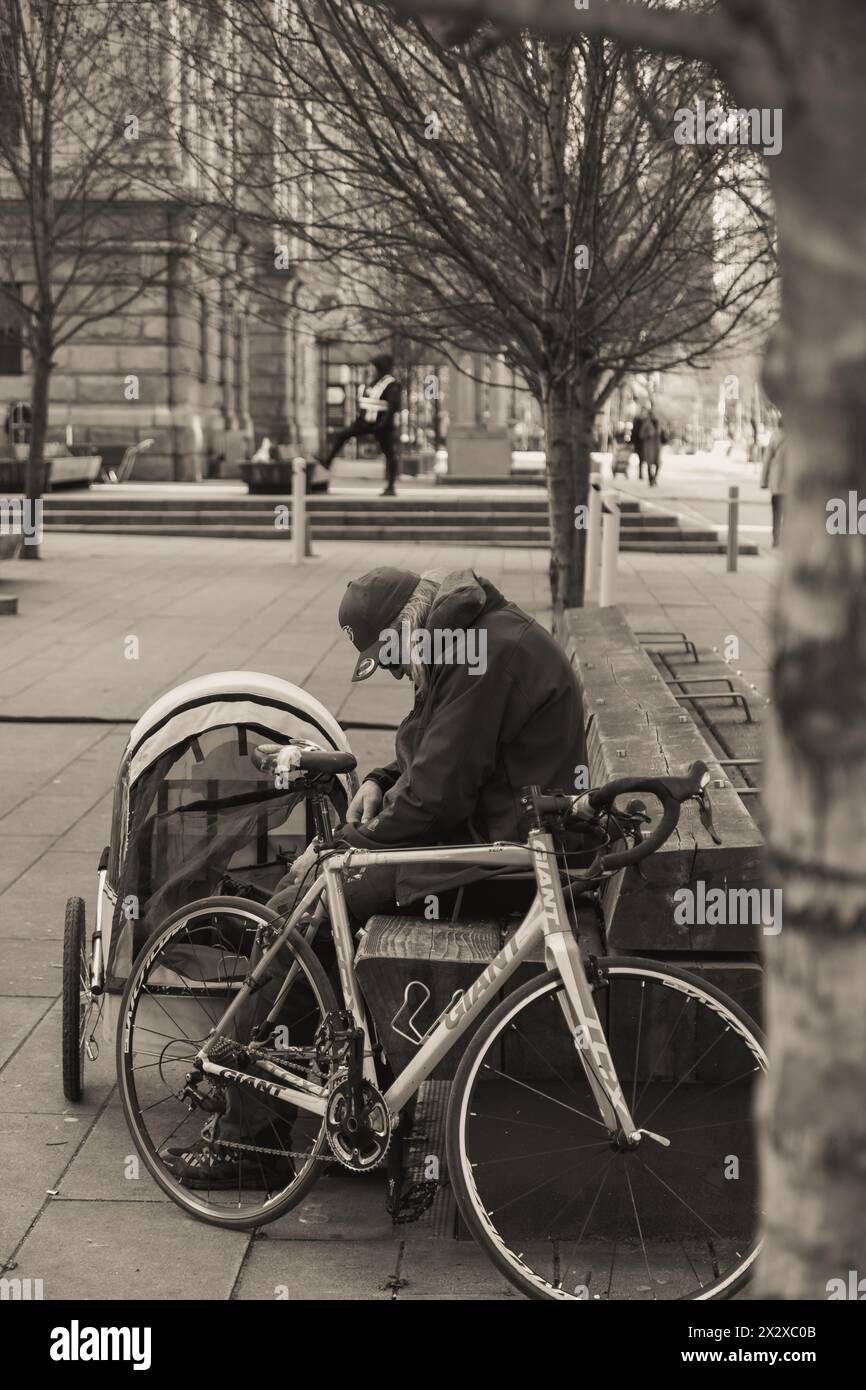 A man sitting on a bench at the Vancouver Art Gallery square, looking at something in his lap, with his bicycle and what appears to be a pet carrier. Stock Photo