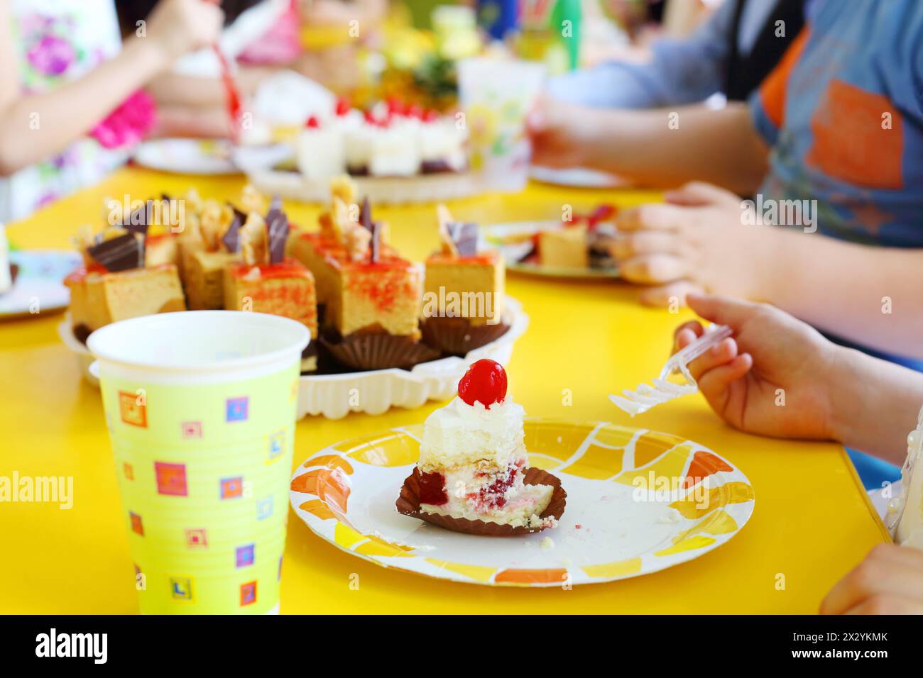 Hands of kids eating delicious little cakes on yellow table. Focus on cake. Stock Photo