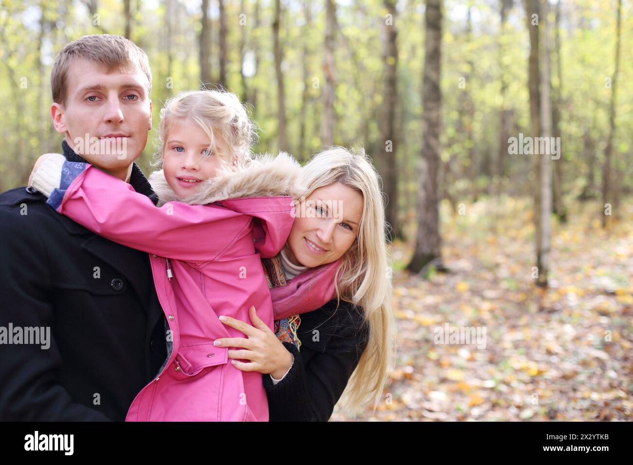 Happy daughter hugs father, and mother peeks out from behind them in autumn forest. Focus on mother. Shallow depth of field. Stock Photo