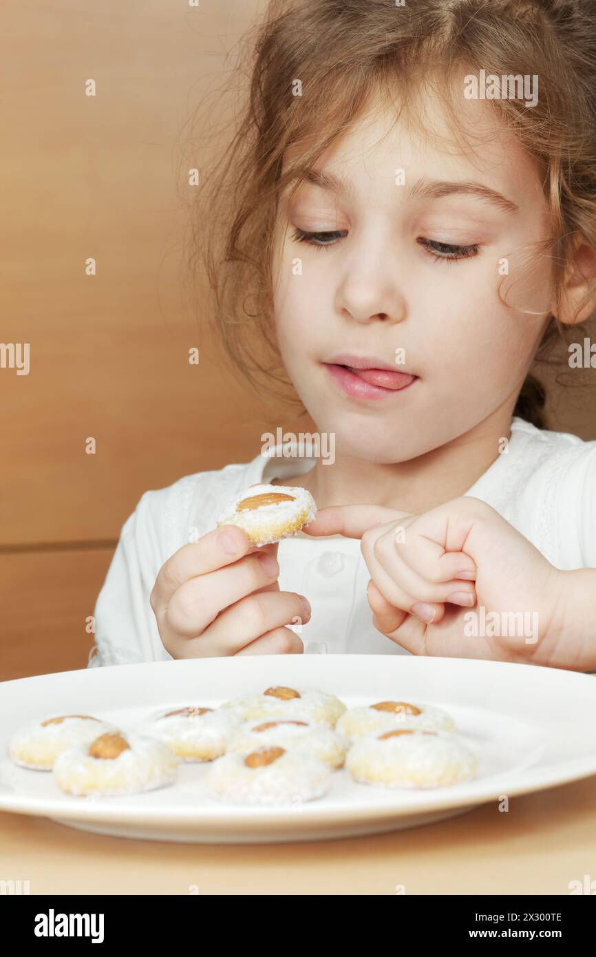 Gir, licking her lips, sits at plate of cookies with almonds and holds one of them Stock Photo