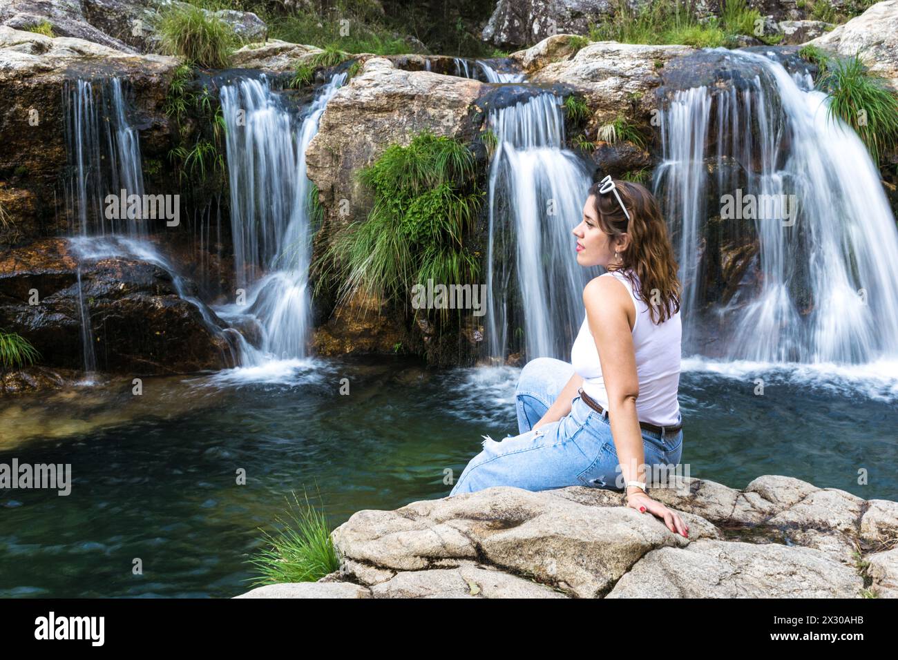 Modern woman sitting on a rock in front of a waterfall in a forest Stock Photo