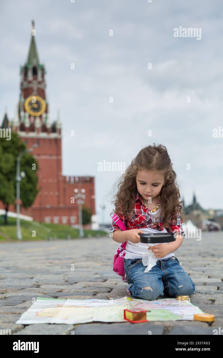 The little girl in stylish dress sitting near the Kremlin on the sett and looking on the map with magnifier Stock Photo