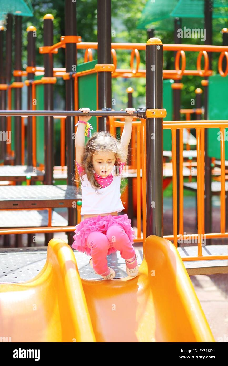 Little girl holds on to the railing to roll down hill on the playground Stock Photo