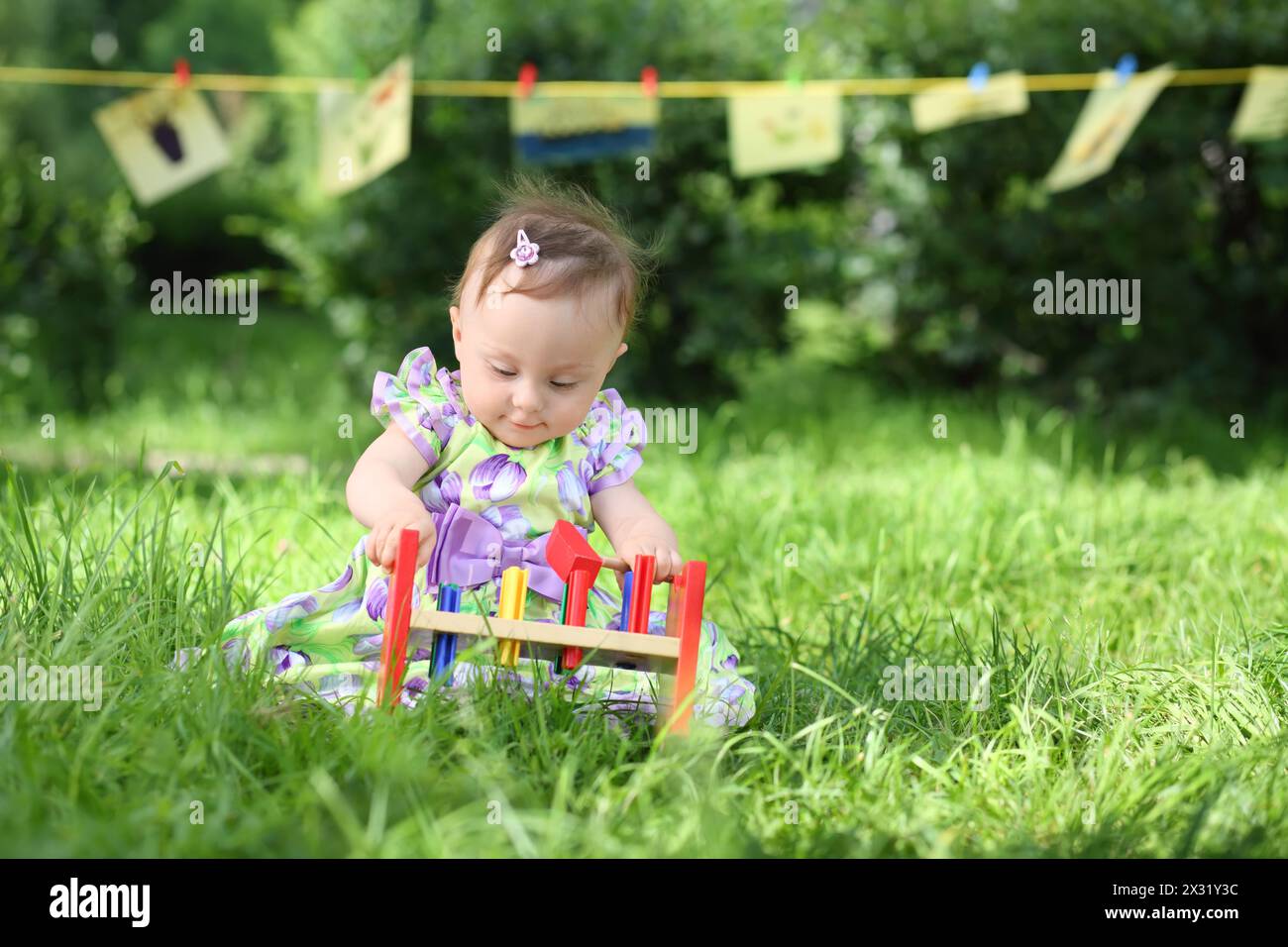 A little girl is playing with colorful wooden toys on the grass, in the background hanging kids drawings Stock Photo