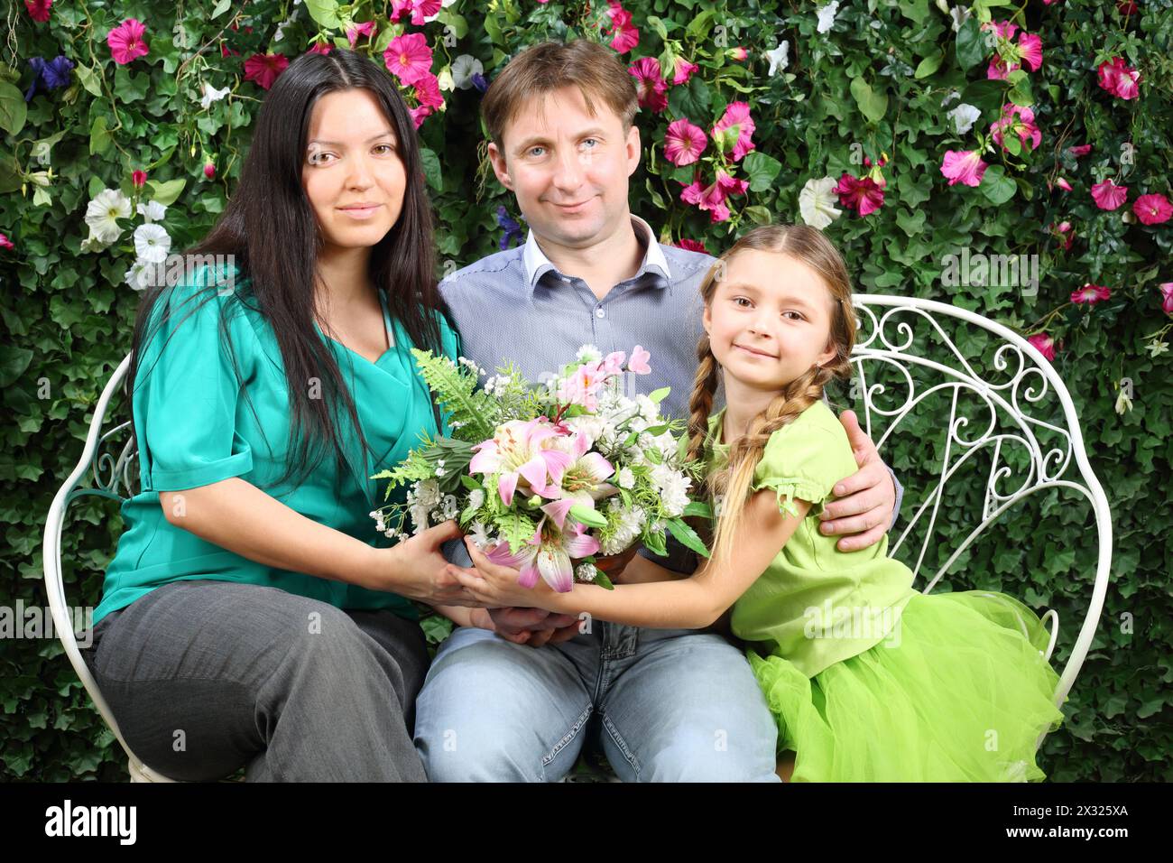 Happy family of three sit on bench, hold flowers and look at camera in garden near verdant hedge. Stock Photo
