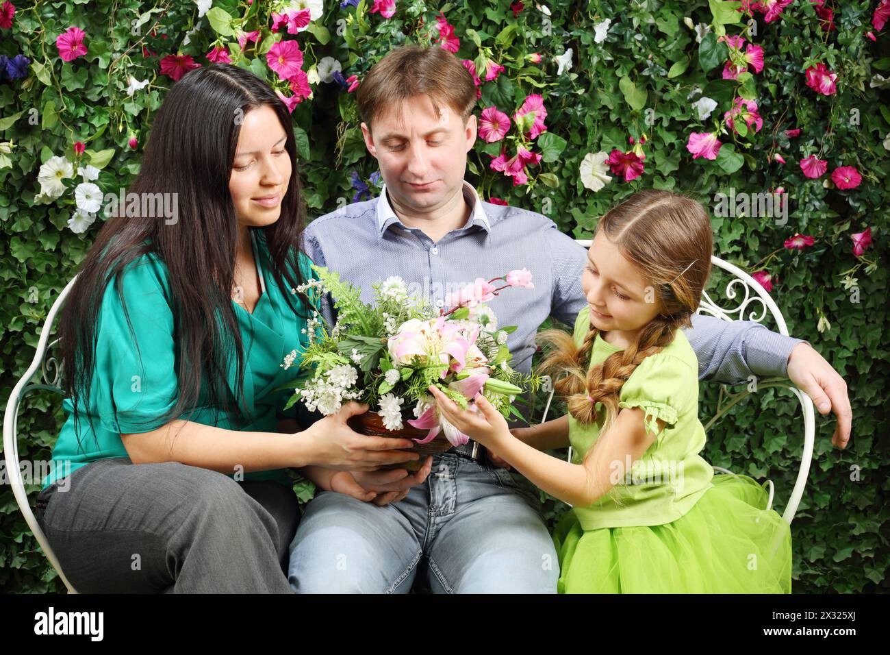 Happy family of three sit on white bench and hold flowers in garden near verdant hedge. Stock Photo