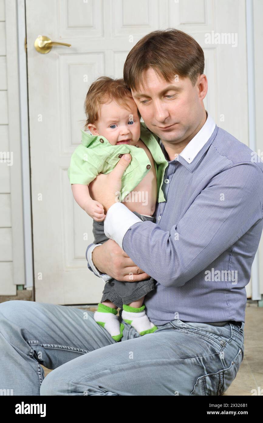 Father holds on hands crying baby and soothes next to white front door. Stock Photo