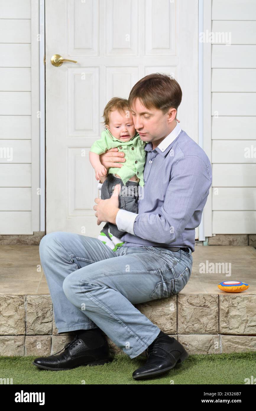 Father holds on hands crying baby, soothes and sits on steps near door. Stock Photo