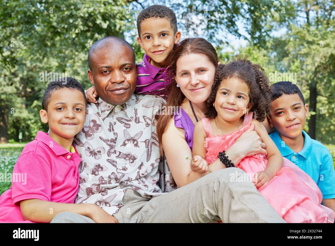 Big interracial family of six sits tight embraced in grass on lawn in park Stock Photo