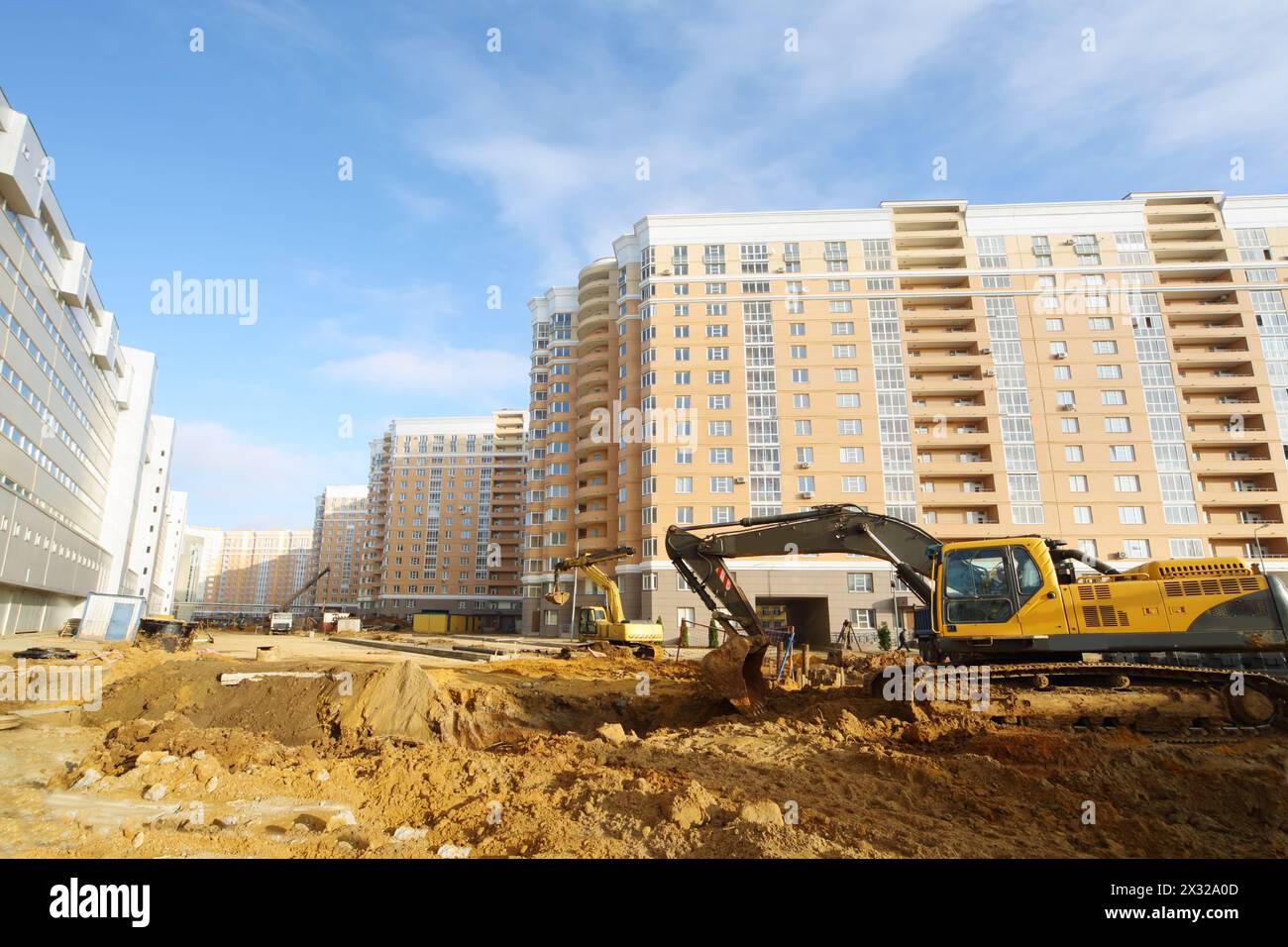 Excavators dig ground near high multi-storey building under construction and parking (on left) at sunny day. Stock Photo