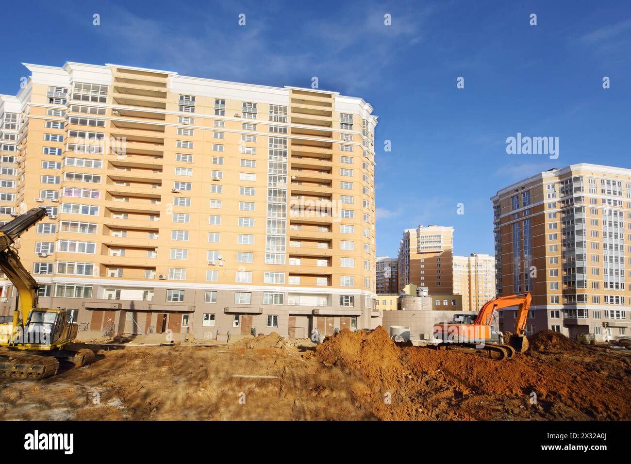 Diggers dig ground near high multi-storey building under construction at sunny day. Stock Photo