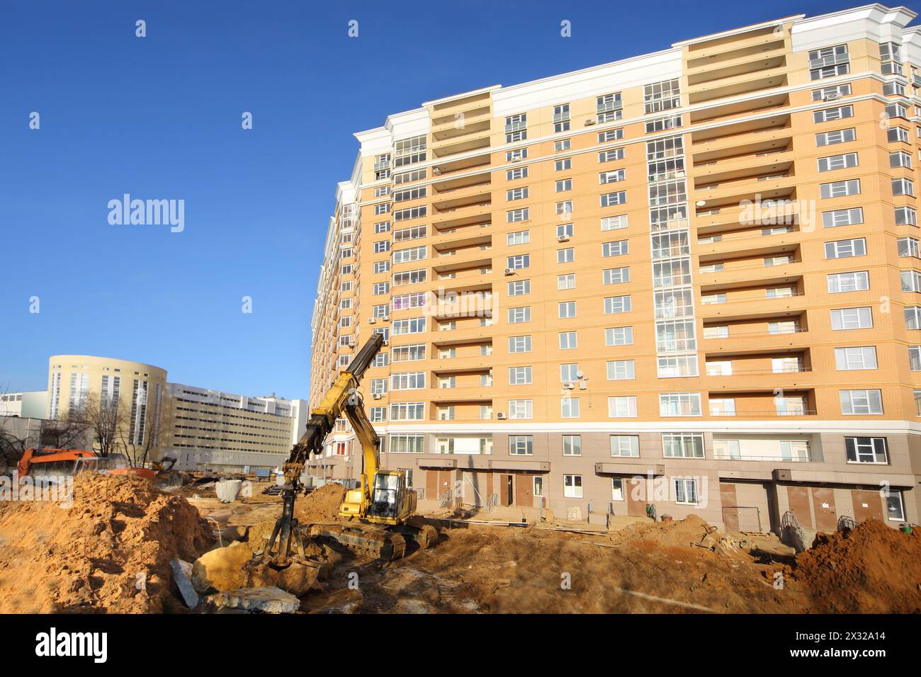 Dredger digs ground near high multi-storey yellow building under construction at sunny day. Stock Photo