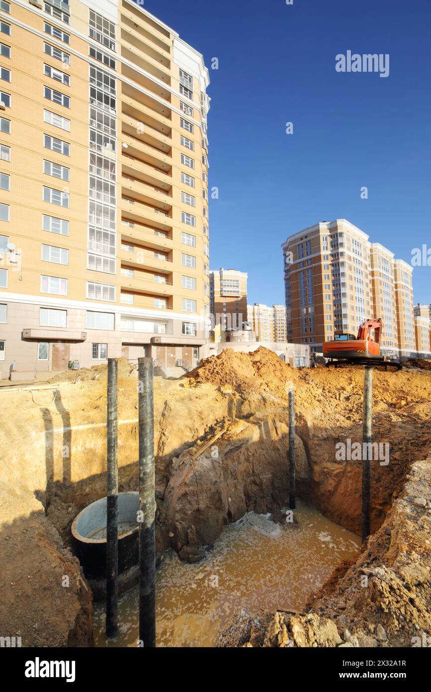 Pit for sewerage with dirty water near high multi-storey yellow building under construction at sunny day. Stock Photo