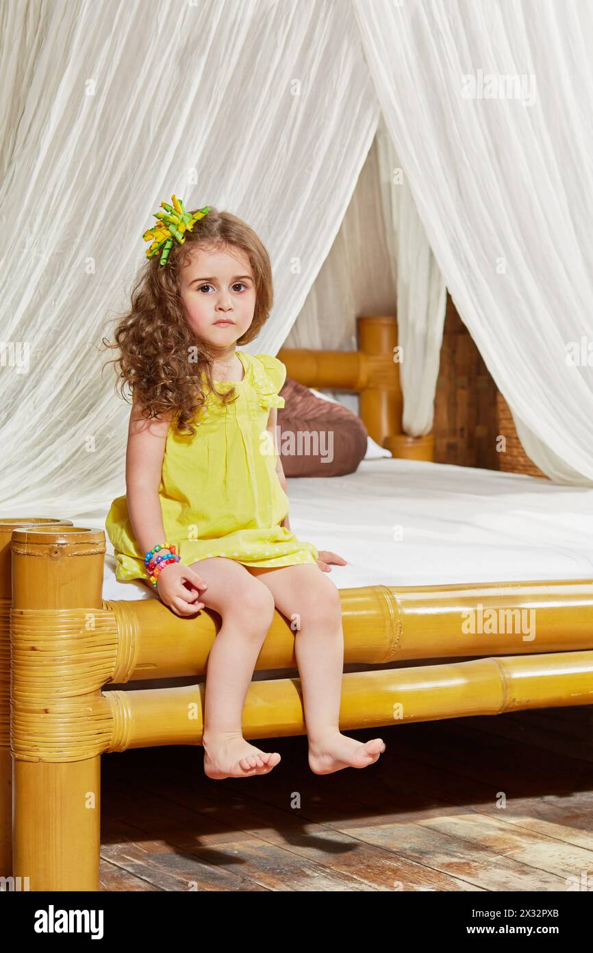 Little girl sits on edge of bed with bamboo frame Stock Photo