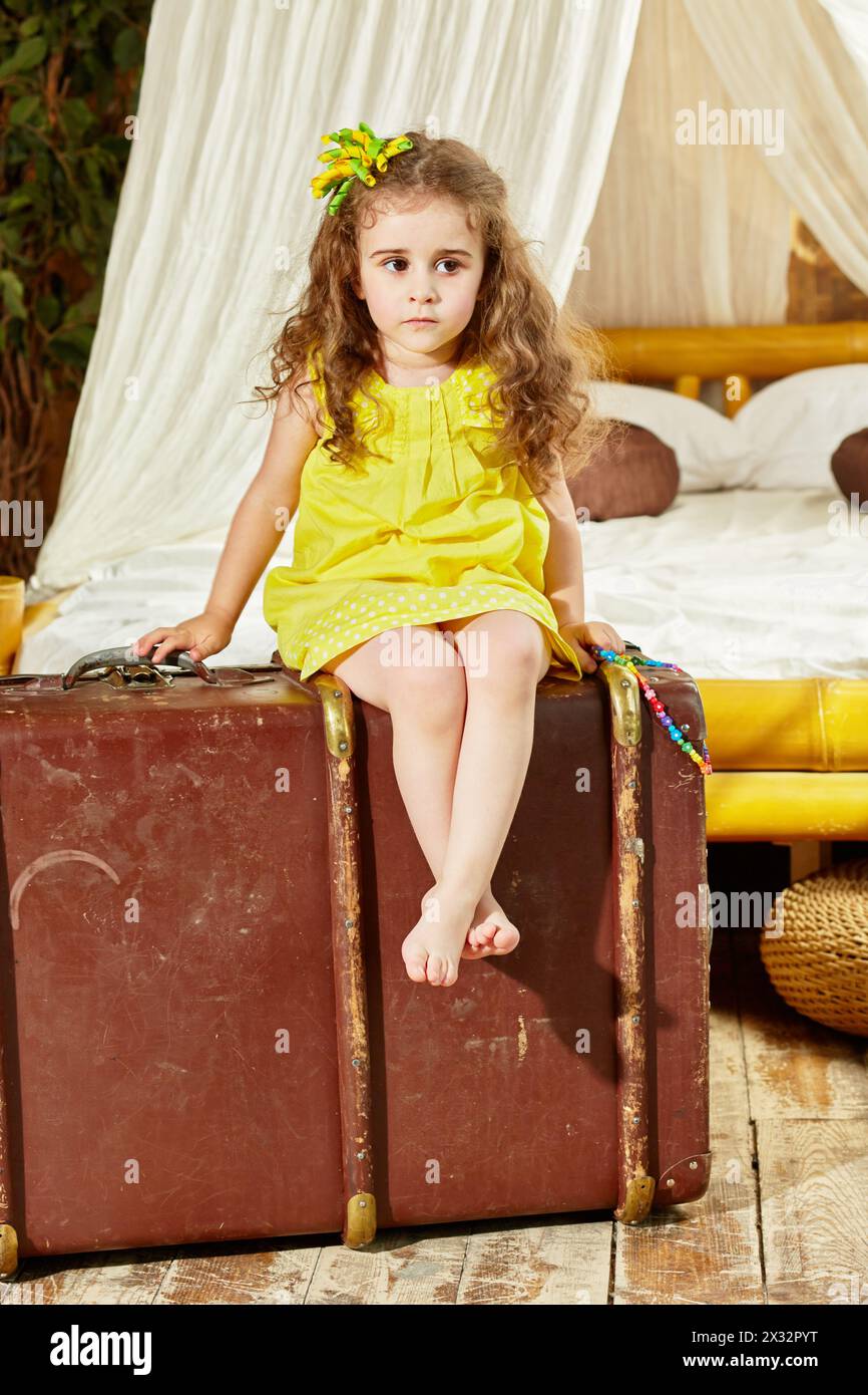 Little girl in yellow dress sits on old big ragged suitcase in room with big bed under tent Stock Photo
