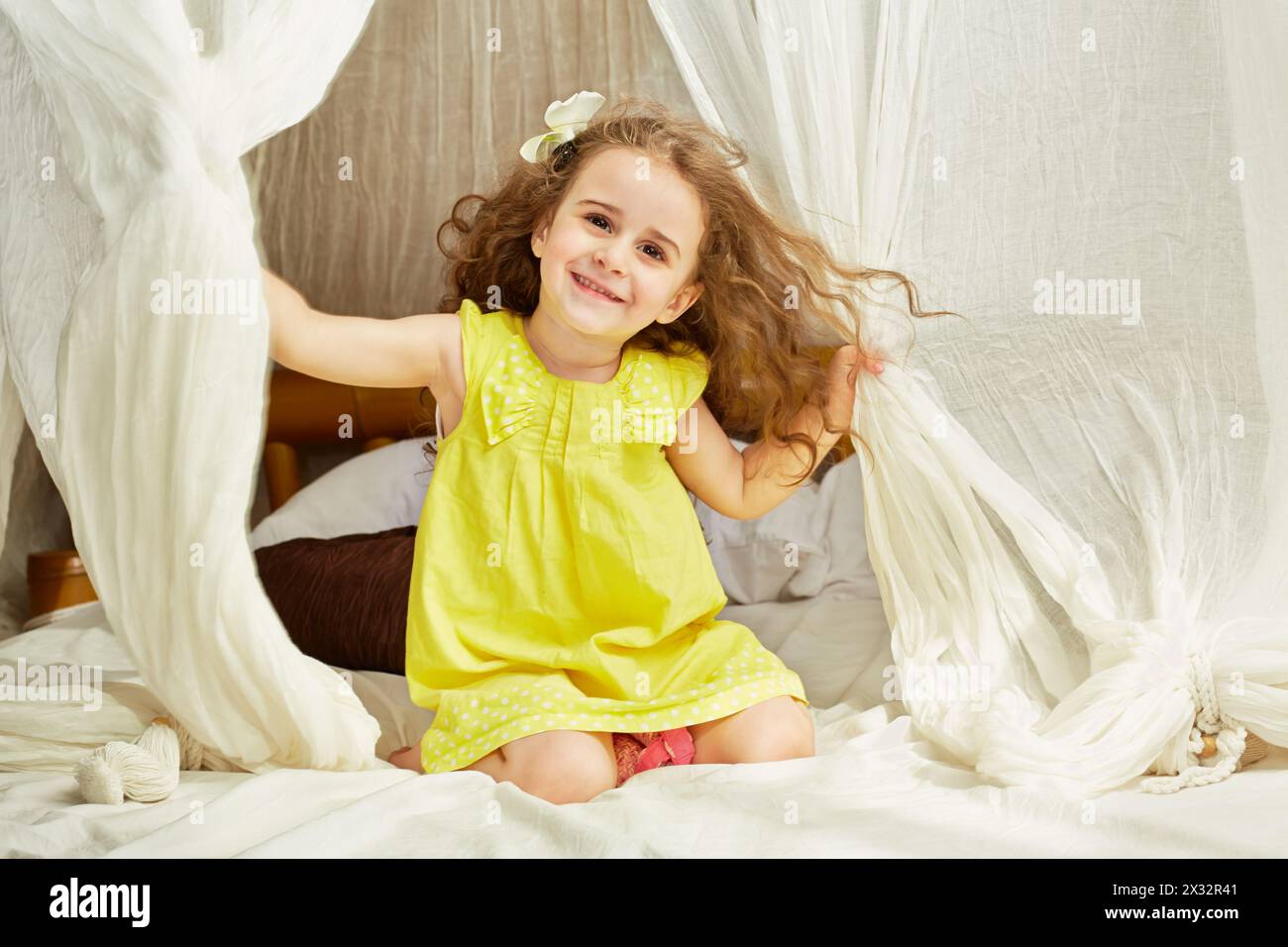 Smiling little girl in yellow dress sits on tent bed Stock Photo