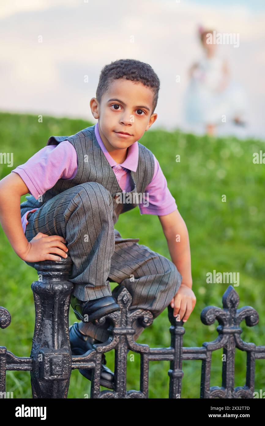 Little boy dressed in suit with vest sits on black wrought-iron fence, figure of little girl behind him not in focus Stock Photo