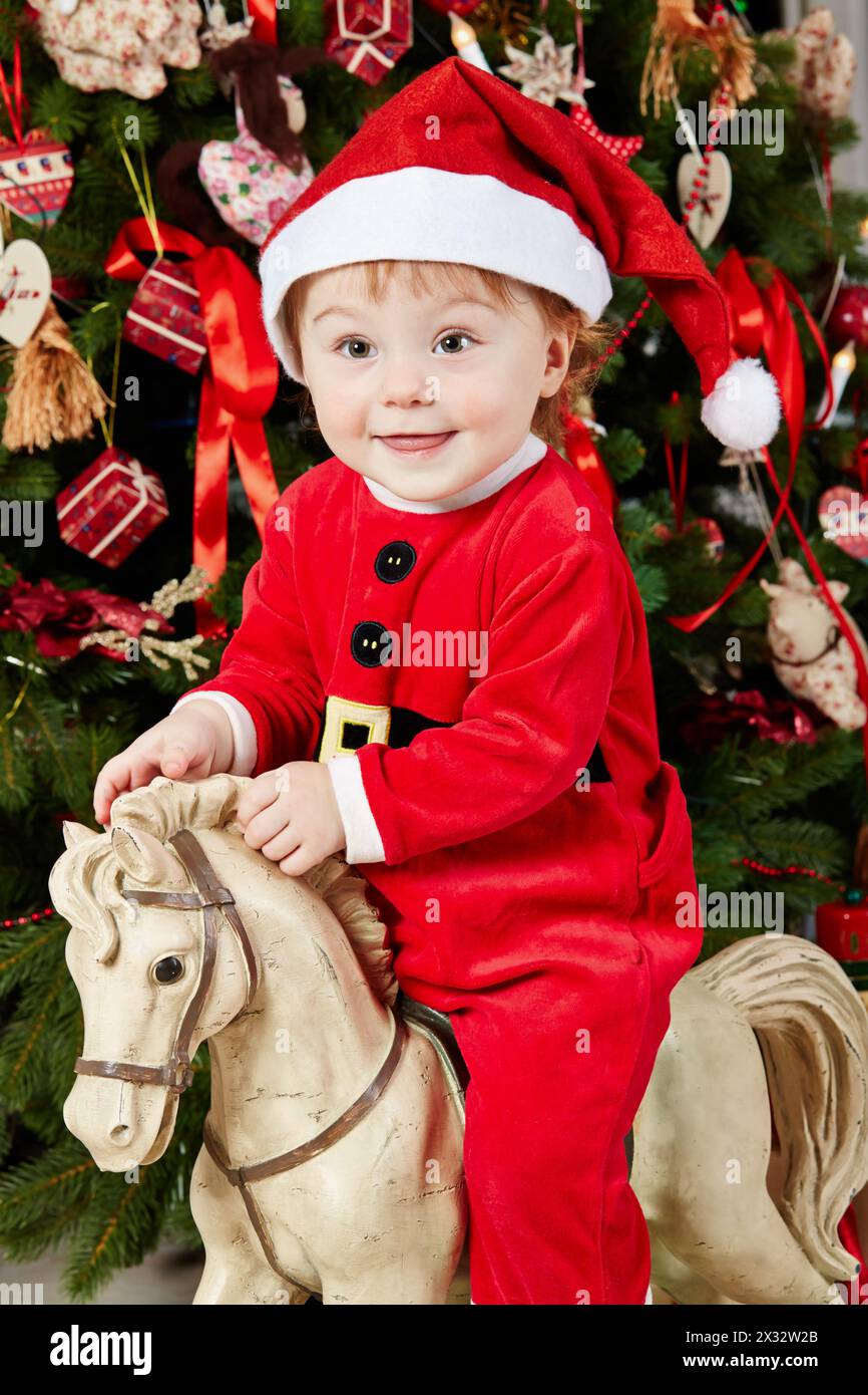Portrait of little girl dressed in santa suit sitting on rocking horse under Christmas tree Stock Photo