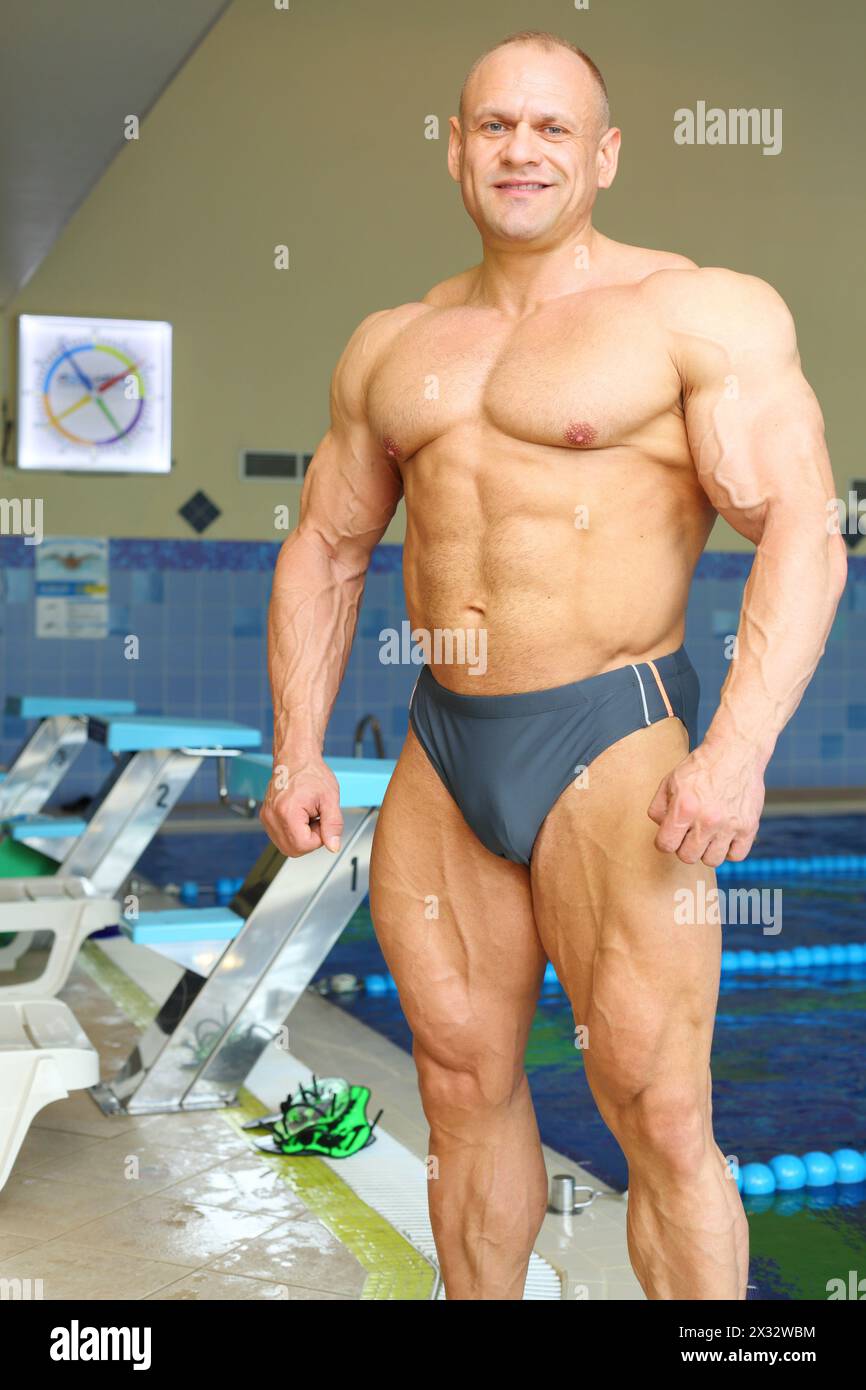 Happy sunburnt bodybuilder stands near indoor pool before swimming in gym hall Stock Photo