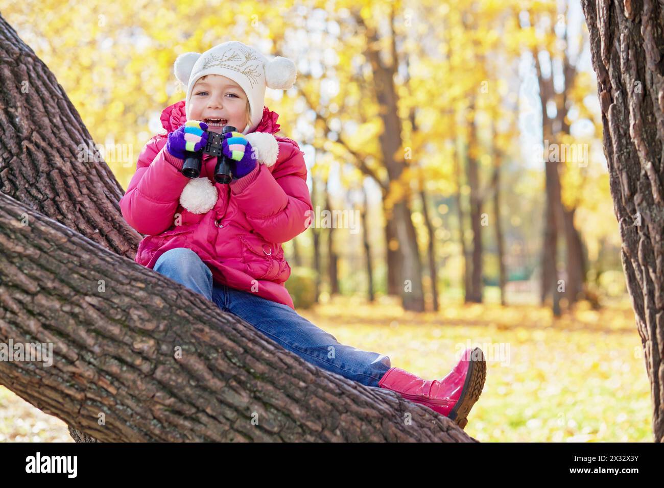 Little girl with binocular sits on inclined tree trunk in autumn park Stock Photo