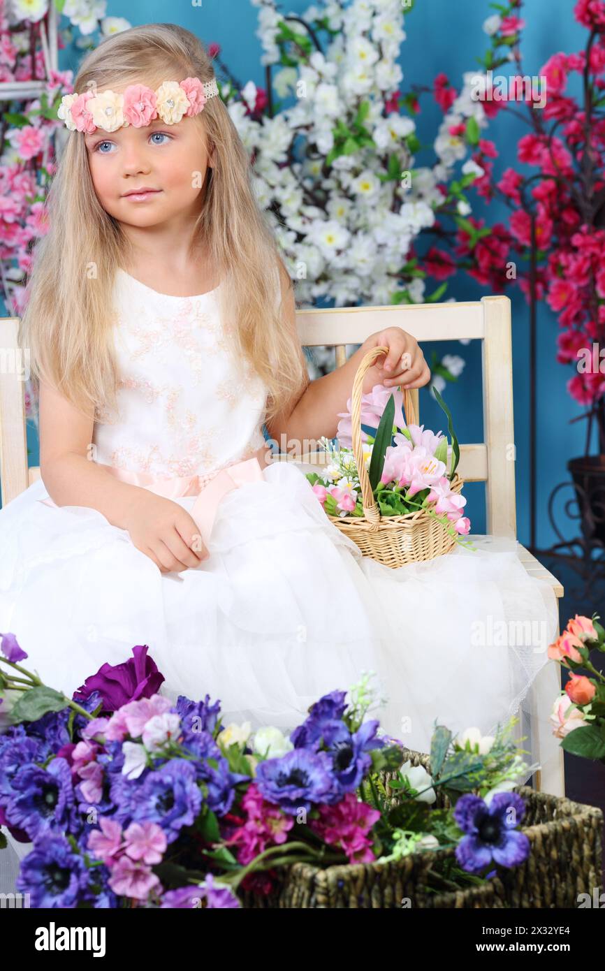 Portrait of blonde little girl in white dress holds basket with flowers among flowers. Stock Photo