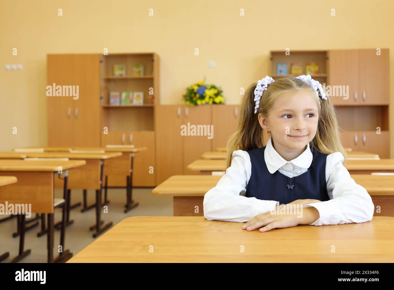 Little girl in school uniform sits at school desk and looks away in classroom in first grade for her alone. Stock Photo